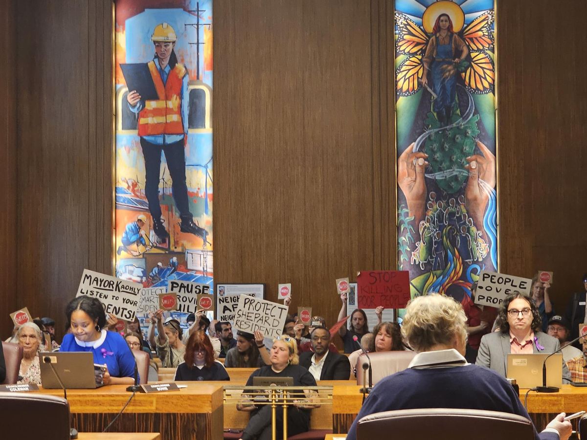 Residents in the Saint Paul City Council chambers hold signs reading Stop the sweeps, Protect Shepard residents, People over property and Mayor Kaohly listen to your residents, as council members sit at the dais.